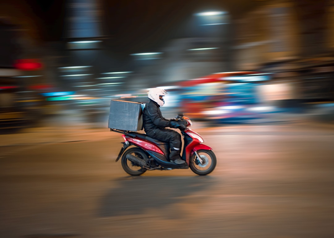 A delivery driver on a red scooter riding through a city at night